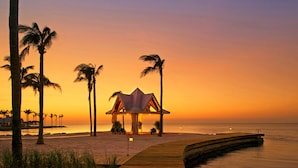Escena de playa con una terraza de madera, palmeras y una estructura tipo pabellón.