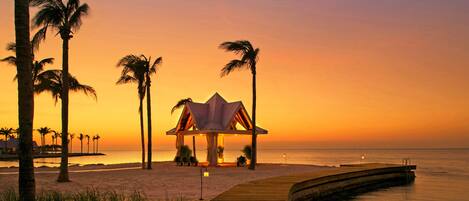Escena de playa con una terraza de madera, palmeras y una estructura tipo pabellón.