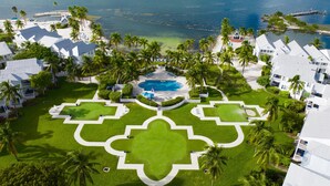 Vista aérea de un resort con una piscina central, rodeado de exuberante vegetación y palmeras, con vista al océano.
