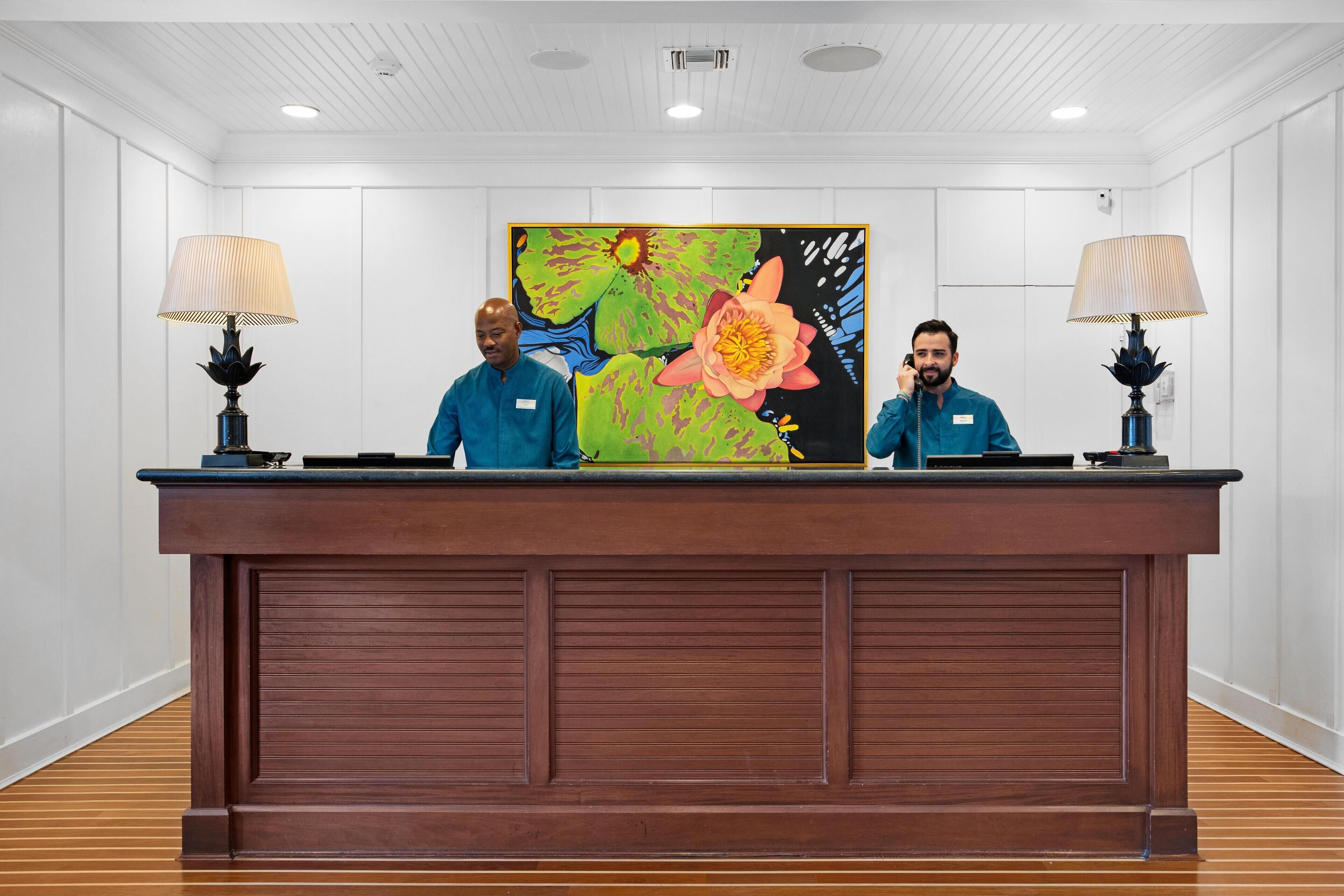 A reception desk with two staff members, a large floral painting, and two lamps.