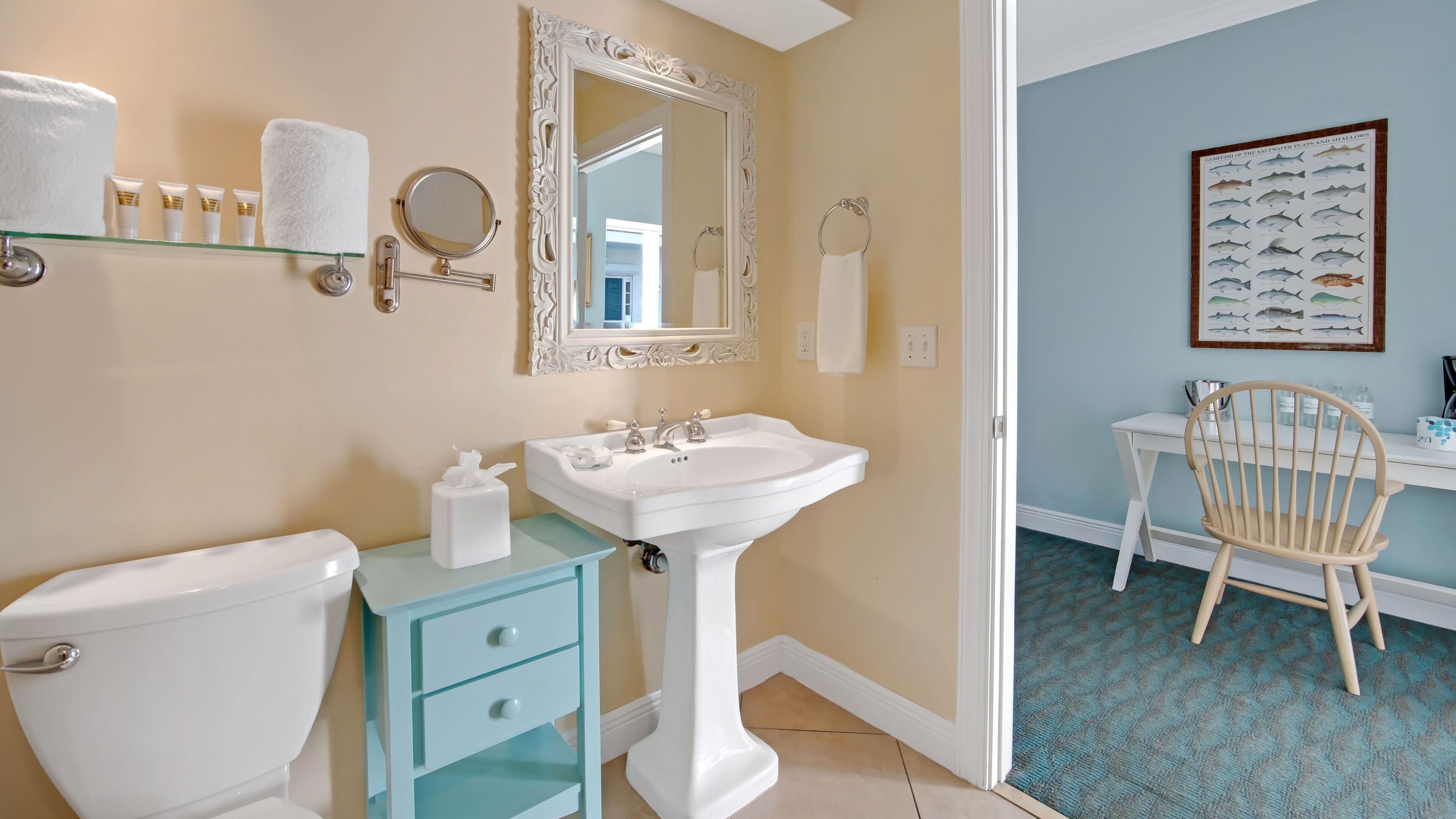 A bathroom with a toilet, sink, and mirror. There is a towel rack with towels and toiletries. A framed fish illustration is on the wall.