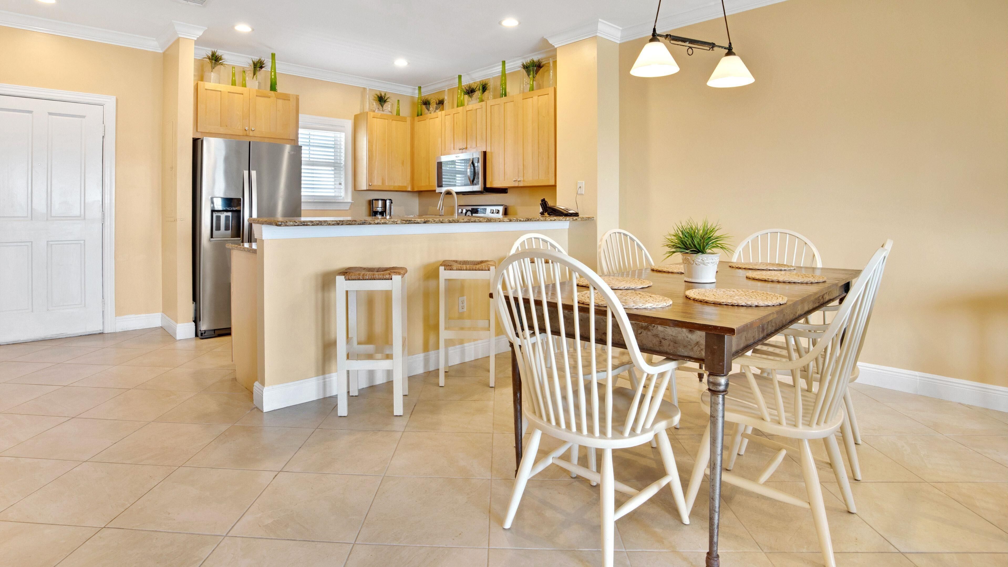 A modern kitchen with wooden cabinets, a stainless steel refrigerator, and a dining area with white chairs and a wooden table.