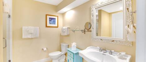 A bathroom with a toilet, sink, and mirror. There are towels hanging on the wall and a framed picture above the toilet.