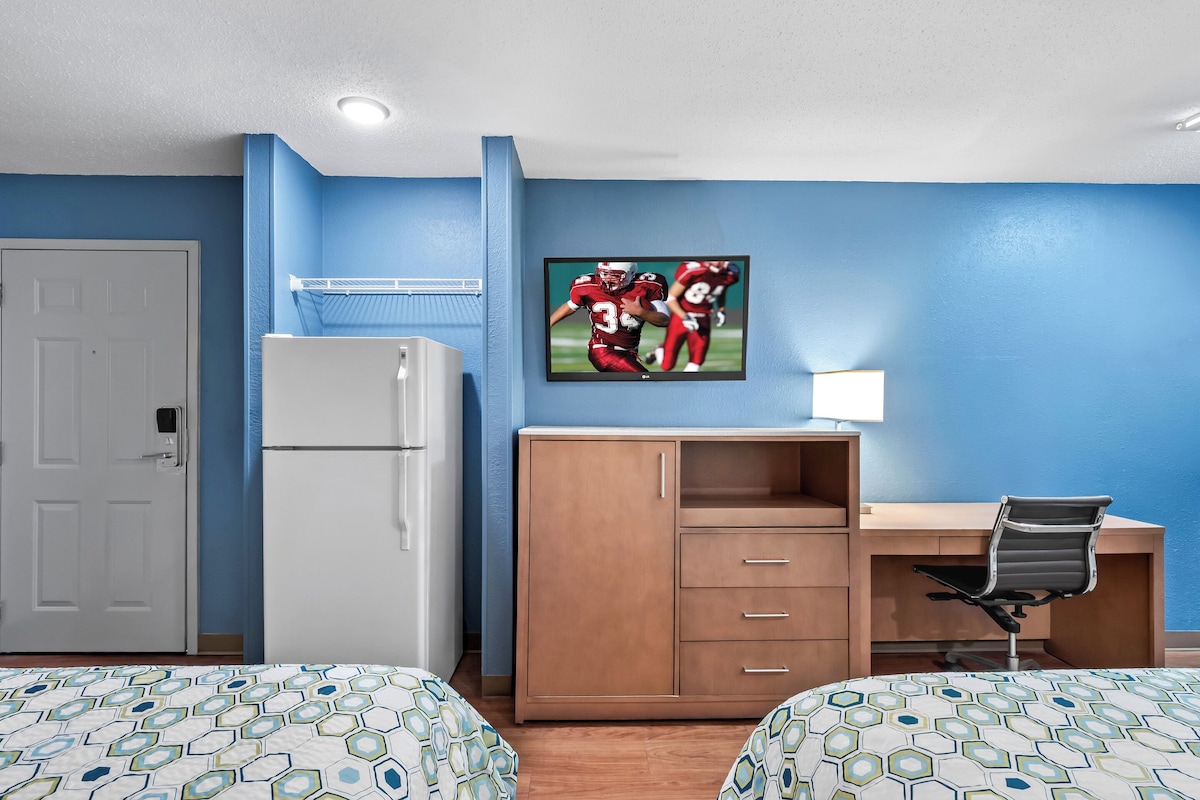 Habitación de hotel con dos camas, un escritorio, un refrigerador y un televisor en la pared.