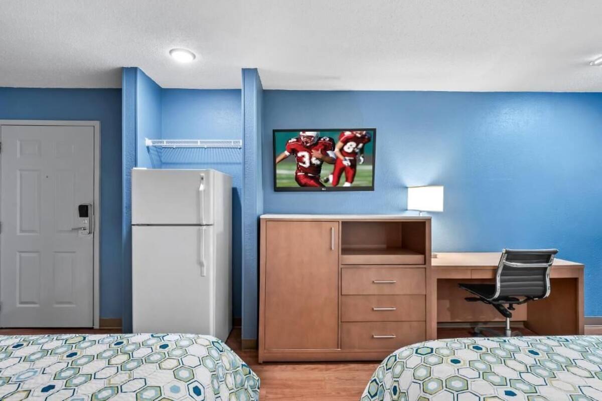 Ein Hotelzimmer mit zwei Betten, einem Schreibtisch, einem Kühlschrank und einem an der Wand befestigten Fernseher.