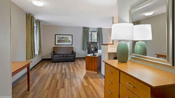 A hotel room with a wooden floor, a sofa, a desk with a lamp, a mirror, and a picture on the wall. - Extended Stay America Suites Philadelphia Airport Bartram Av (Philadelphia)