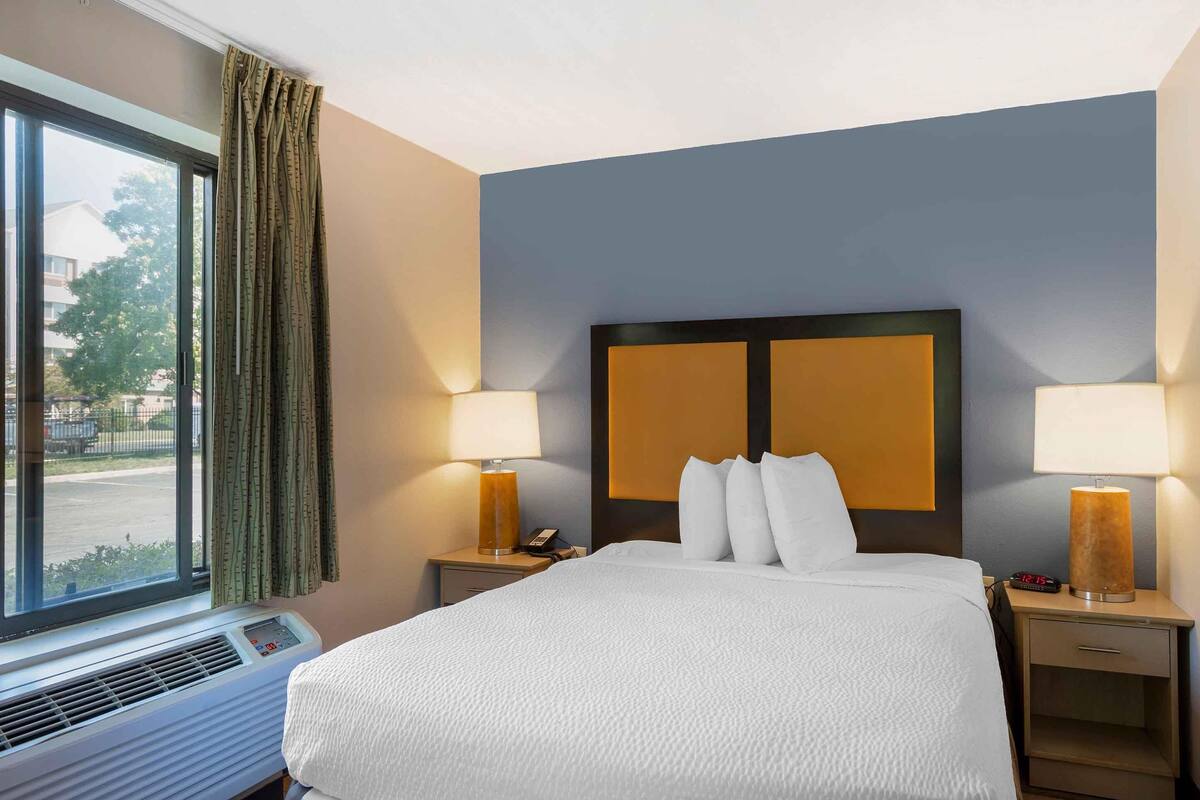 Een hotelkamer met een groot bed, twee bedlampjes, een nachtkastje met een telefoon en een raam met gordijnen.