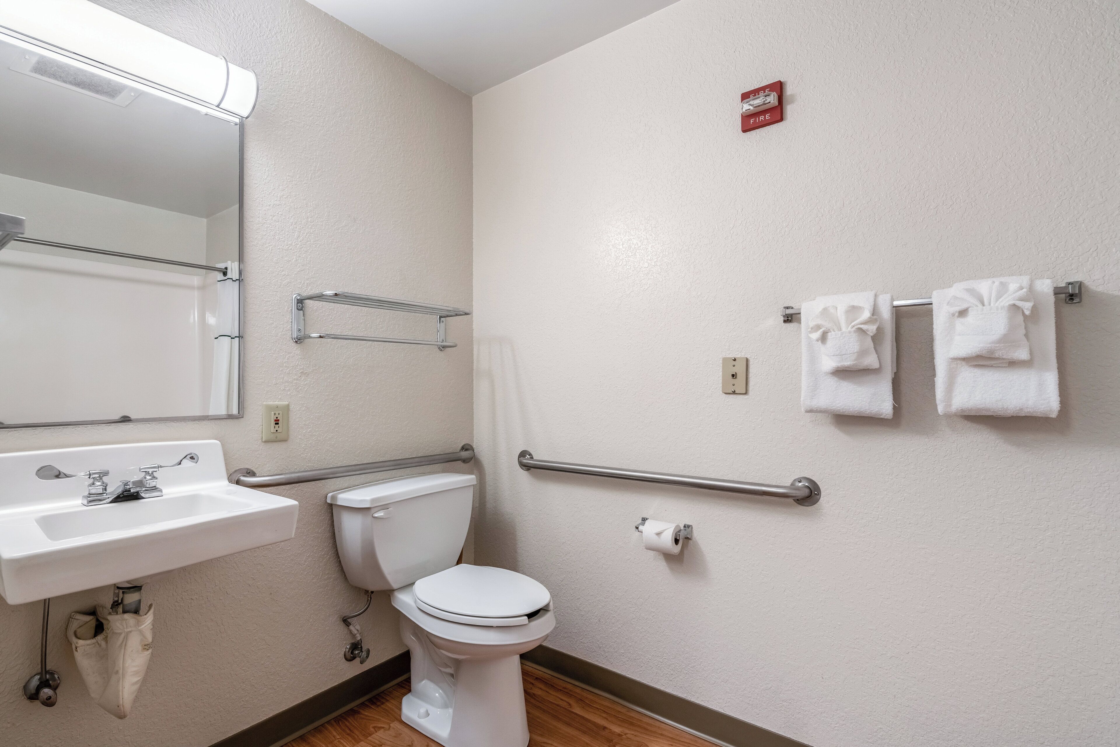 A bathroom with a white sink, toilet, and handrails. There is a towel rack with towels and a fire alarm on the wall.