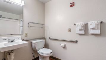 A bathroom with a white sink, toilet, and handrails. There is a towel rack with towels and a fire alarm on the wall.