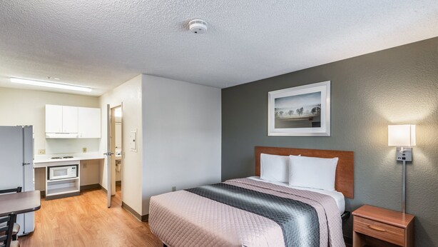 A hotel room with a bed, bedside table, and a small desk with a chair. - HomeTowne Studios by Red Roof Spokane - Valley (Spokane Valley)