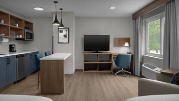 Moderni hotellihuone, jossa on pieni keittiö, taulutelevisio, työpöytä tuolilla ja suuri ikkuna, jossa on verhot.