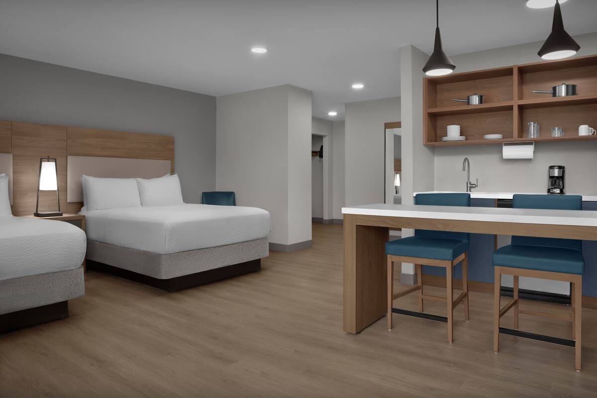A modern hotel room with two beds, a wooden headboard, a dining area with a table and chairs, and built-in shelves with kitchen items.