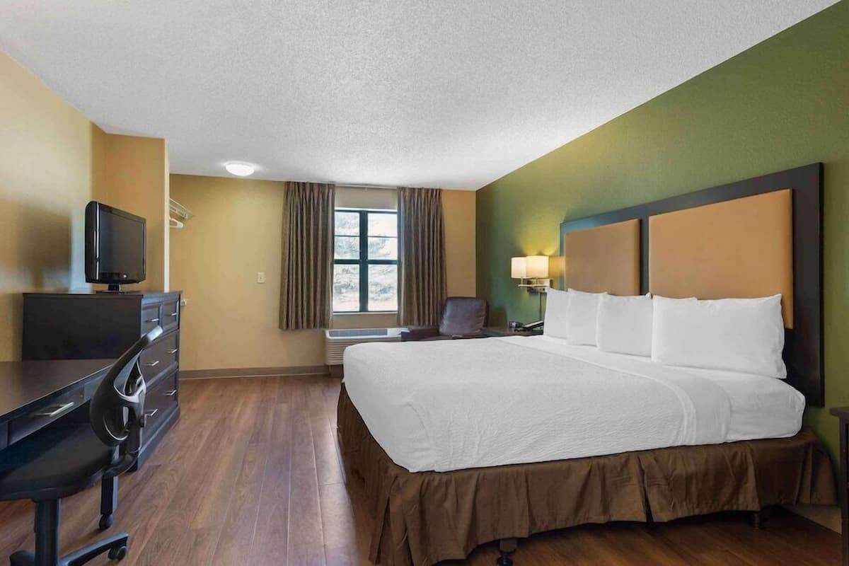 A hotel room with a bed, a desk with a television, a chair, a window with curtains, and a lamp.