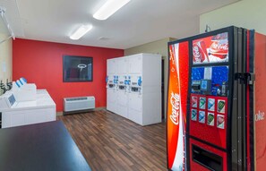Une pièce avec un mur rouge, des casiers blancs, un distributeur automatique de Coca-Cola et une horloge numérique.