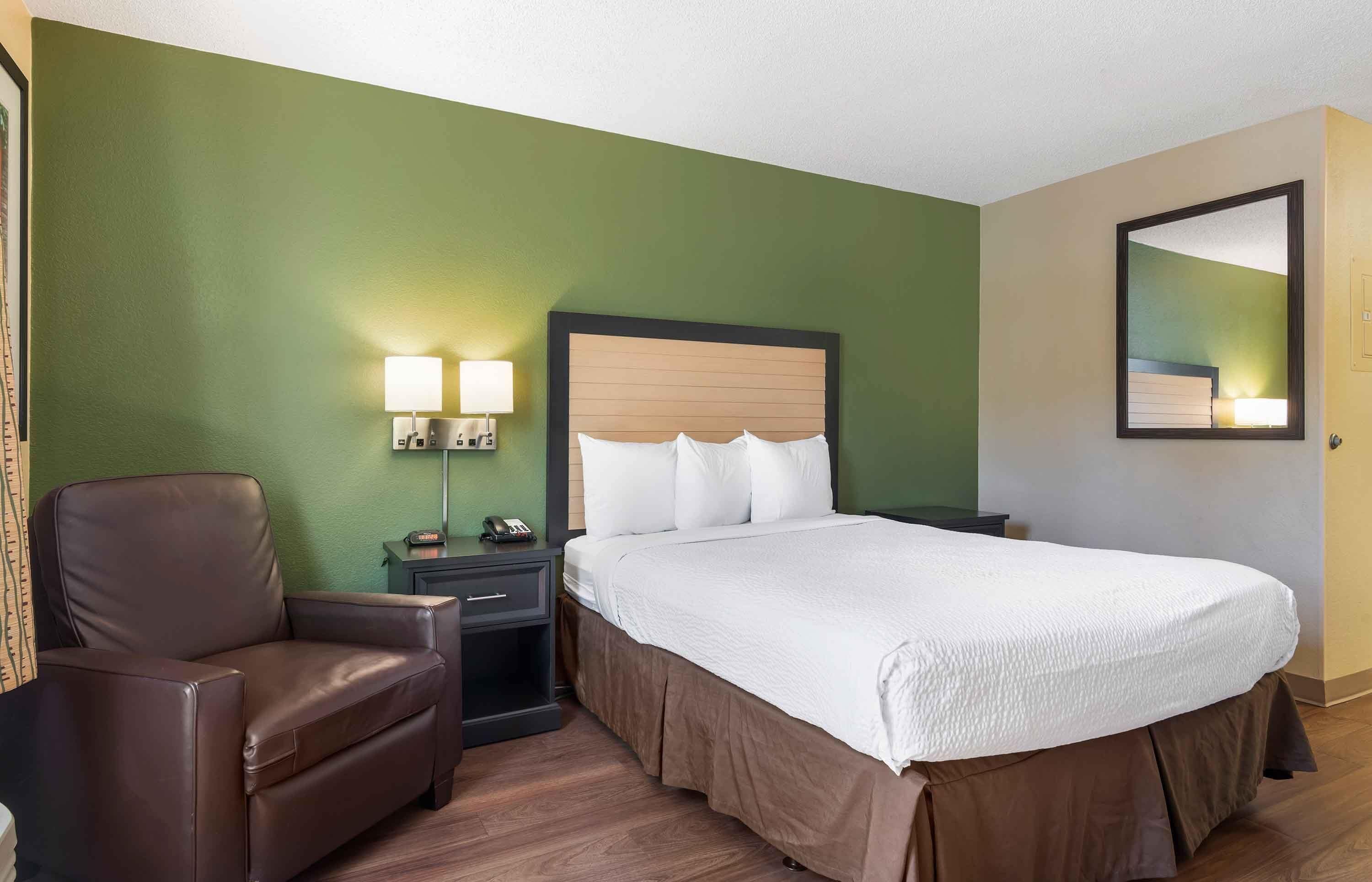 Une chambre d’hôtel avec un lit, une chaise, une table de chevet avec des lampes et un miroir au mur.