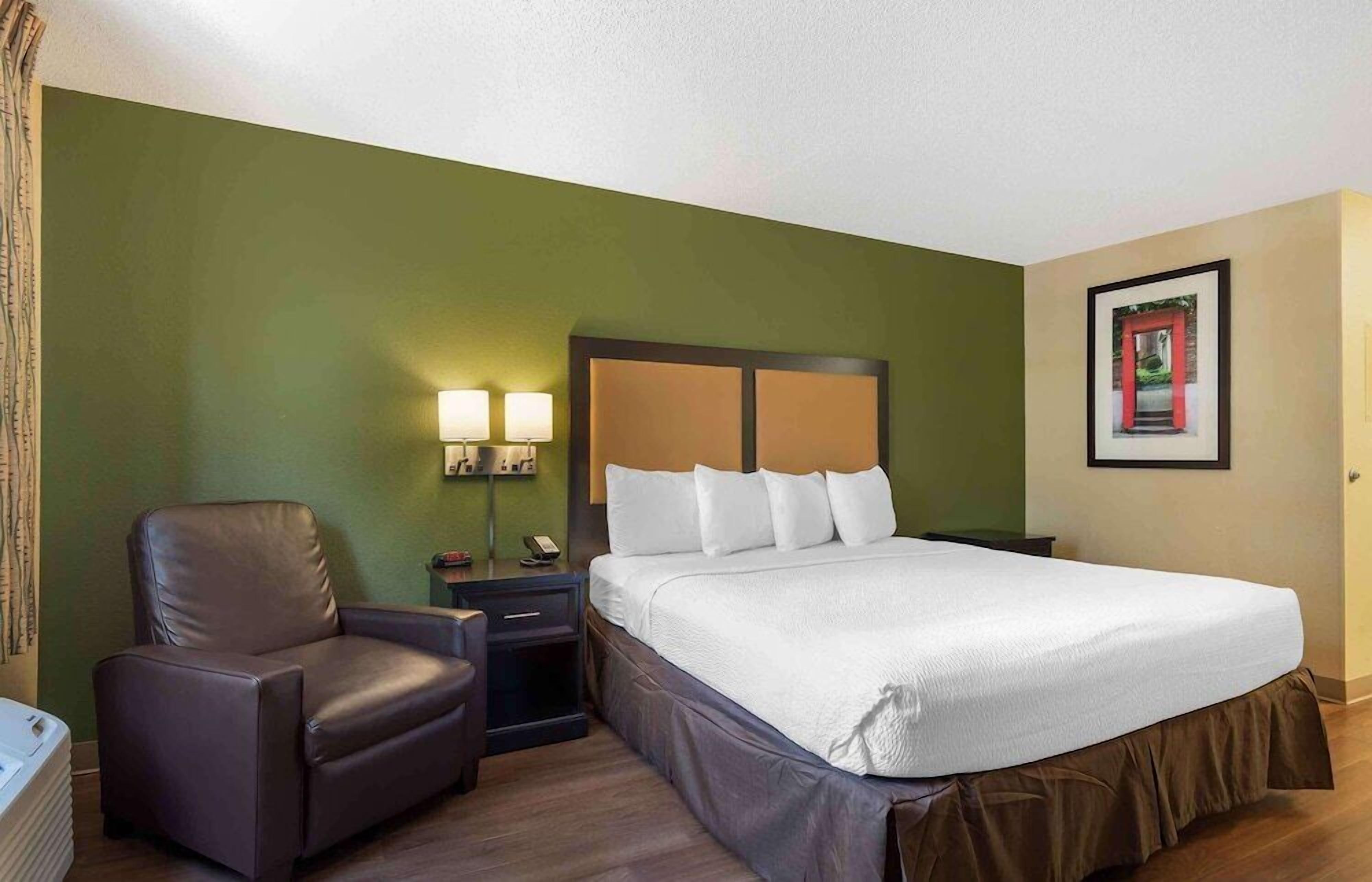 Quarto de hotel com cama, uma cadeira, mesa de cabeceira e quadro na parede.