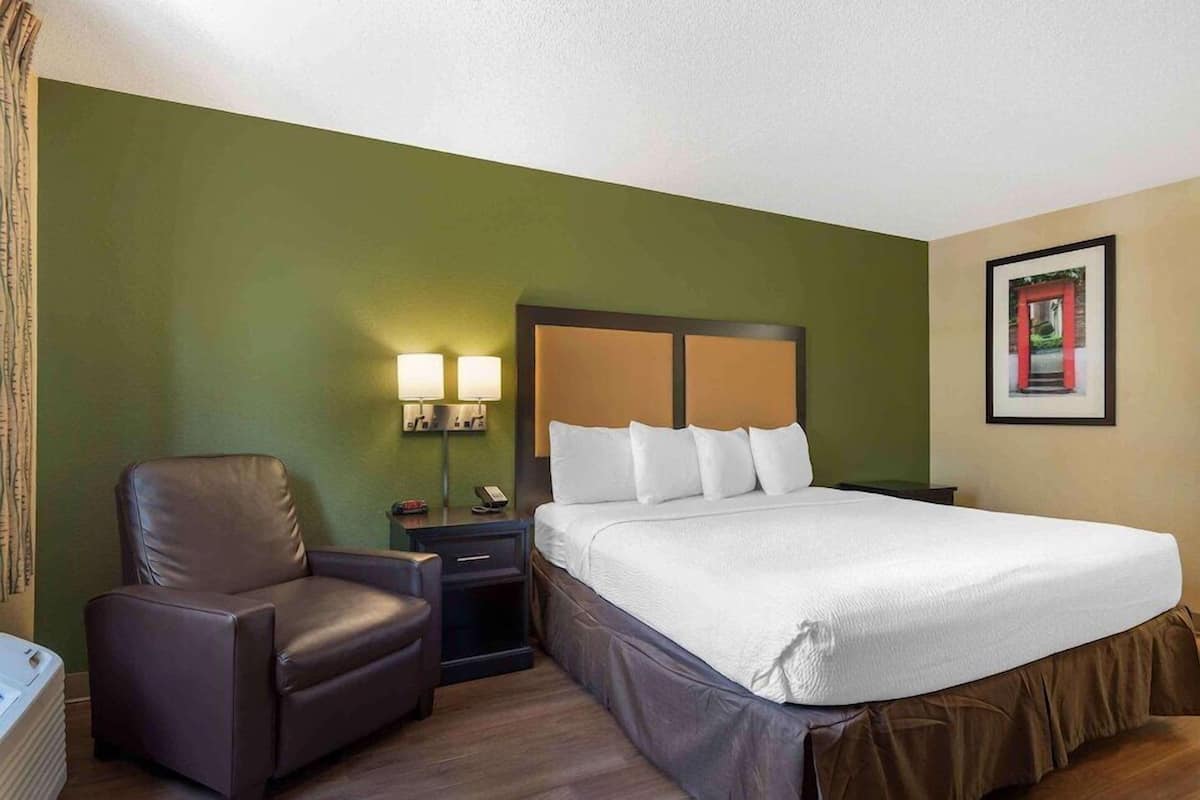 A hotel room with a bed, a chair, a nightstand, and a wall-mounted picture.
