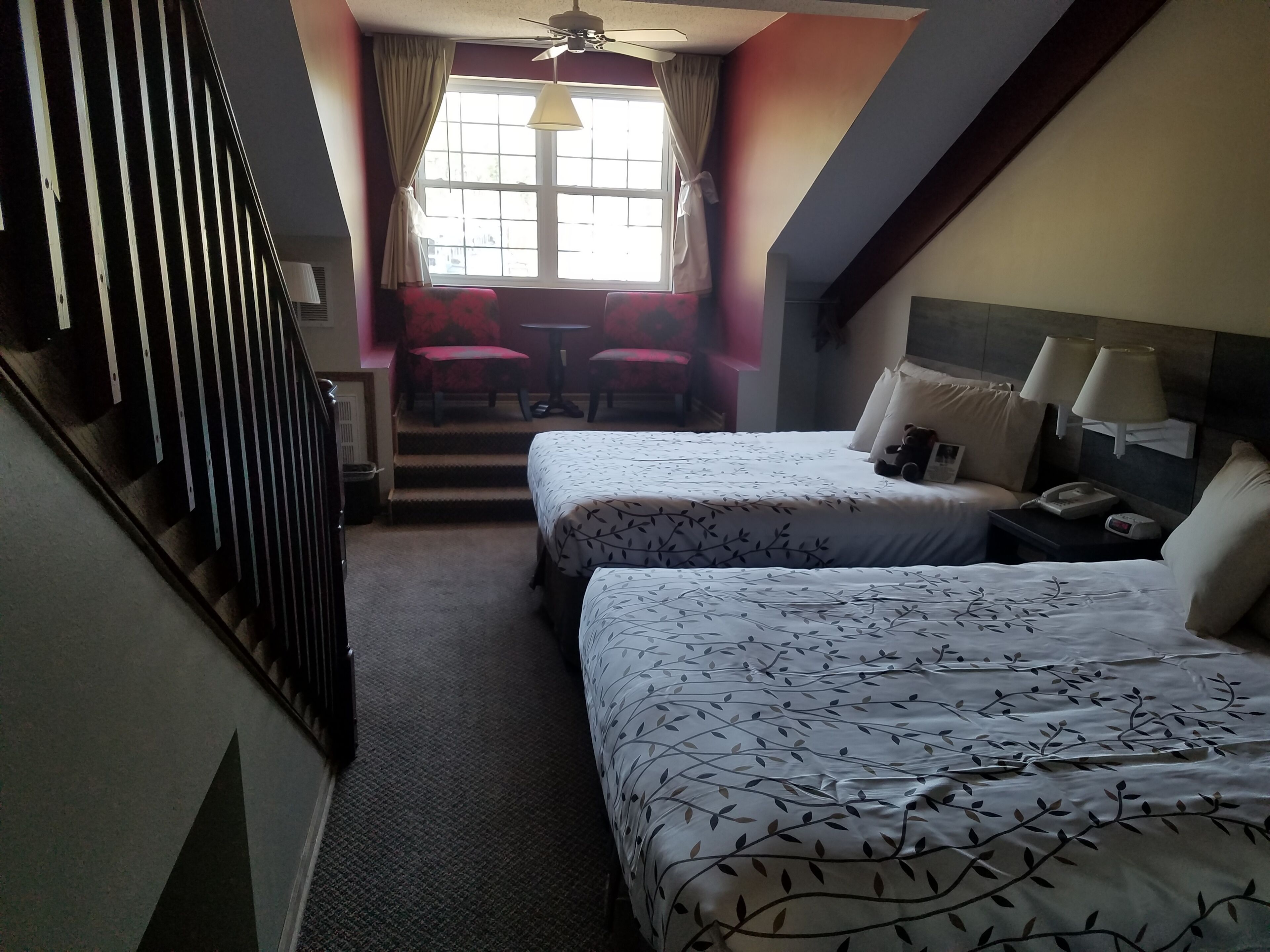 Una habitación de hotel con dos camas, una escalera, una ventana con cortinas, una mesa con sillas y una mesita de noche con lámpara.