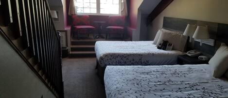 Una camera d'albergo con due letti, una scala, una finestra con tende, un tavolo con sedie e un comodino con una lampada.