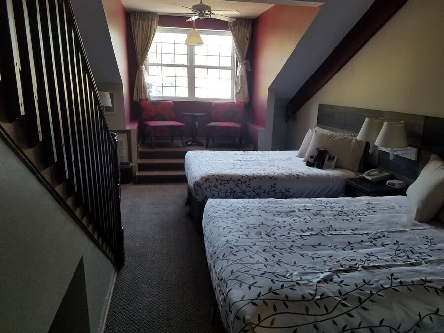 Una habitación de hotel con dos camas, una escalera, una ventana con cortinas, una mesa con sillas y una mesita de noche con lámpara.