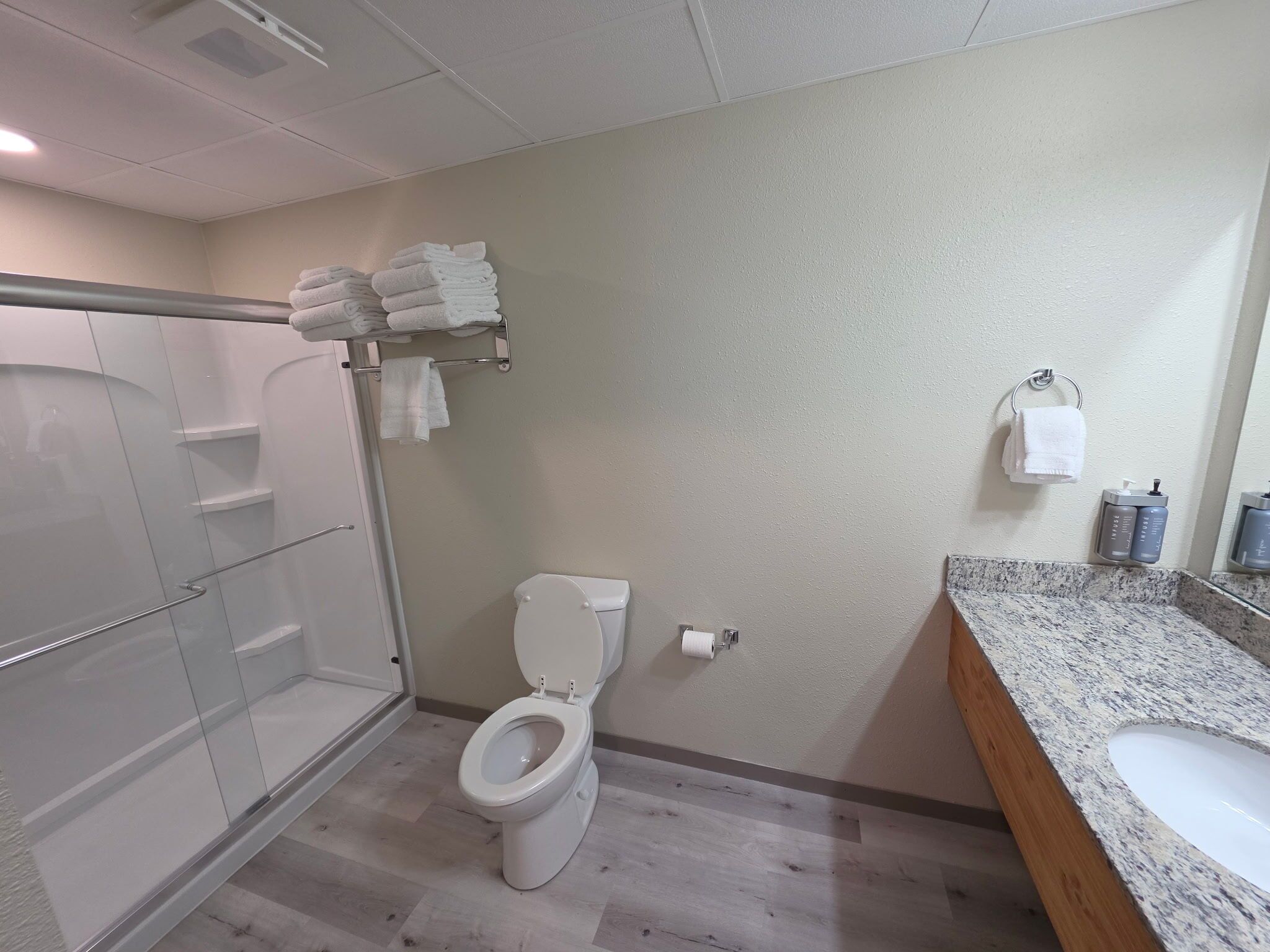 A bathroom with a glass shower enclosure, a toilet, a sink with a granite countertop, and a towel rack with folded towels.