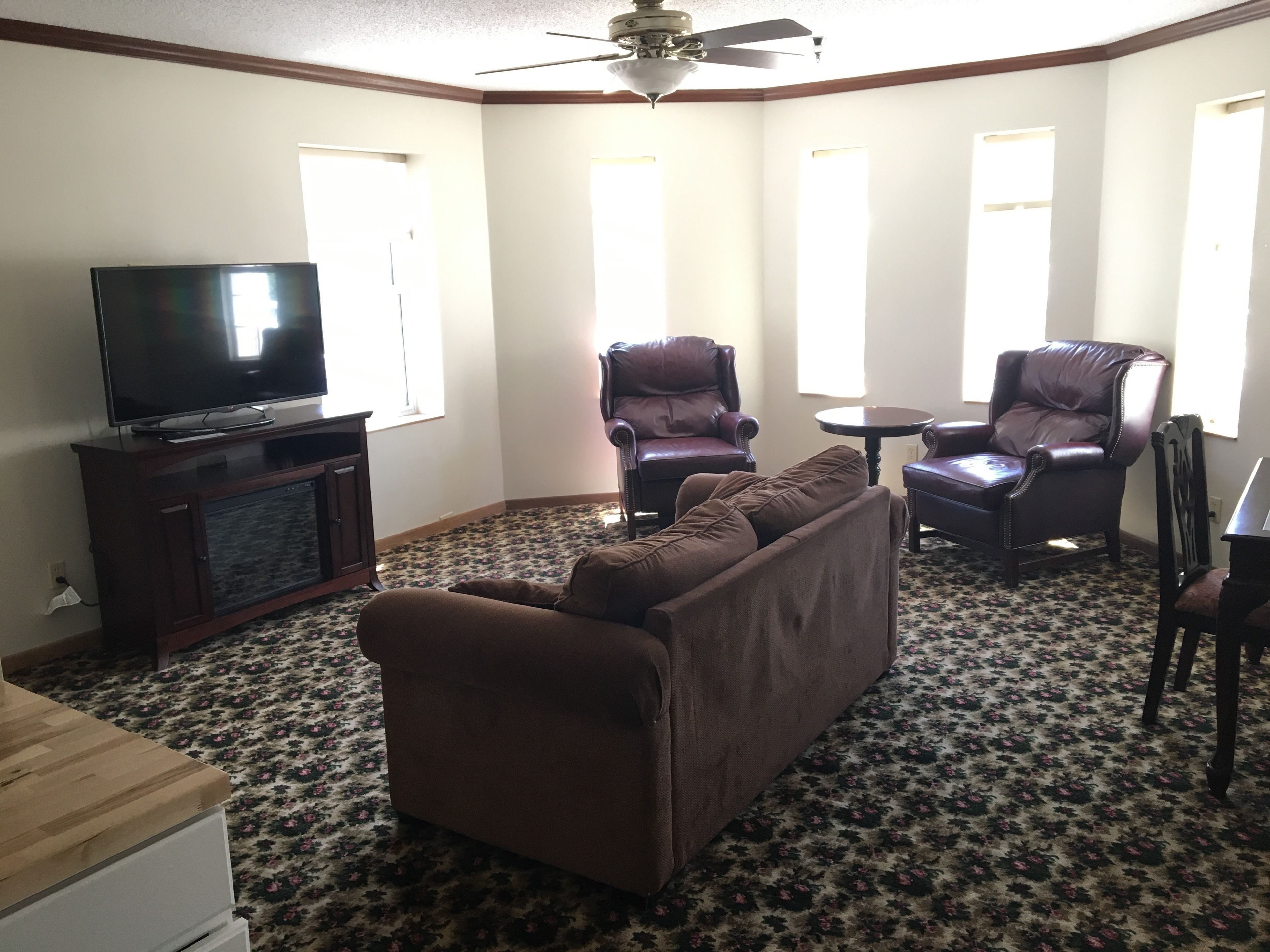 A living room with a brown sofa, two leather armchairs, a small round table, and a television on a wooden cabinet.