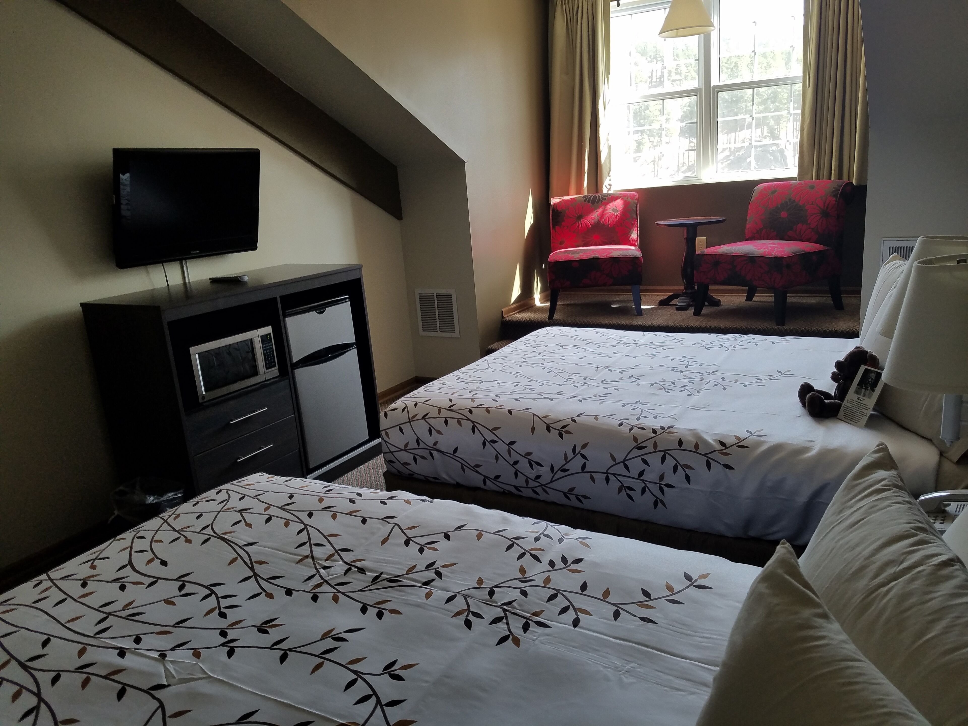 A hotel room with two beds, a TV on a dresser, a window with curtains, and a small table with a chair.
