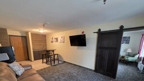 Una sala de estar con un sofá de cuero marrón, una mesita baja con taburetes, un televisor de pantalla plana montado en la pared y una puerta corrediza que da a otra habitación.
