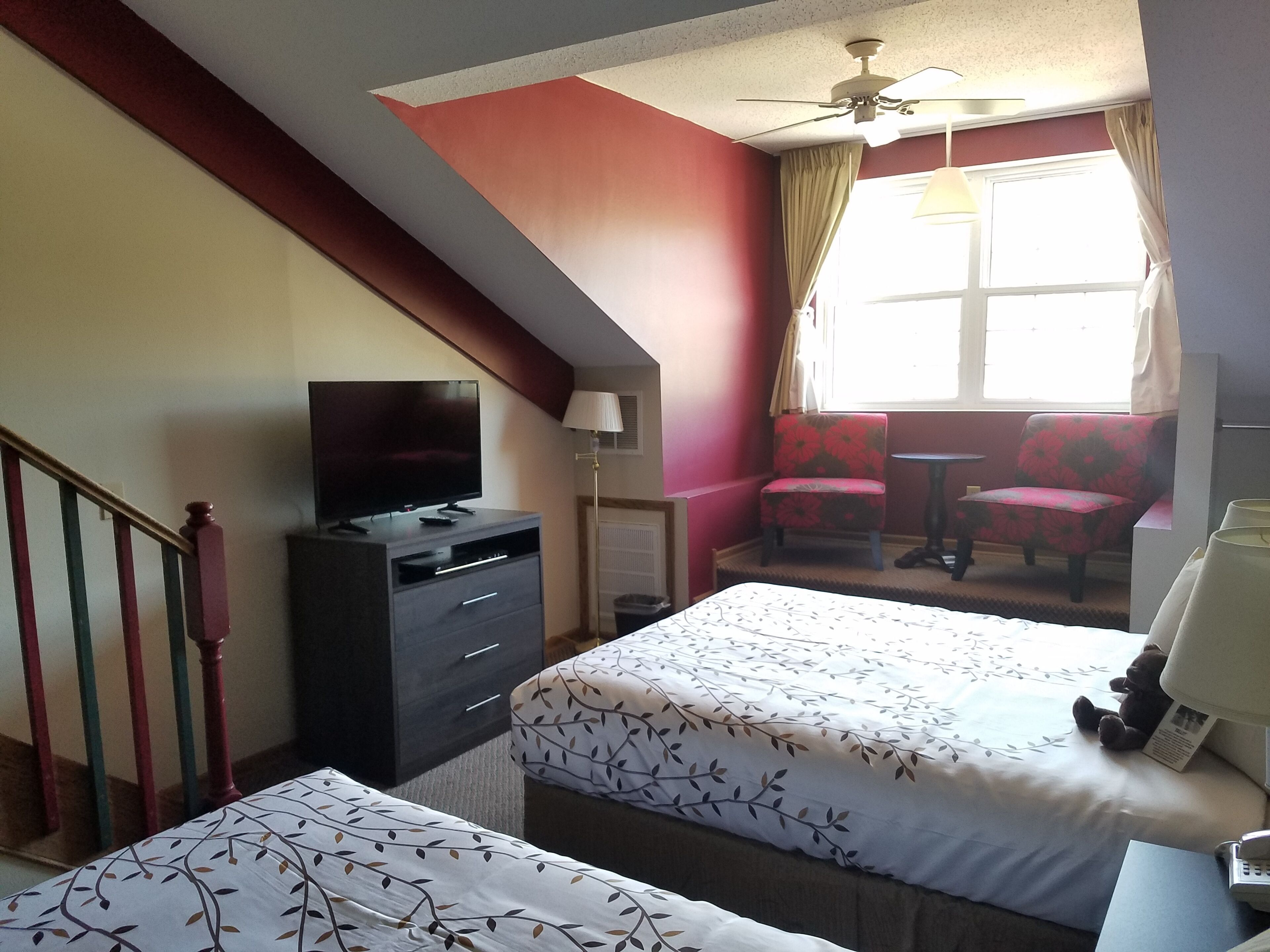 Ein Hotelzimmer mit Bett, Fernseher, Schreibtisch, Stuhl und einem fensterbepflanzten Fenster.