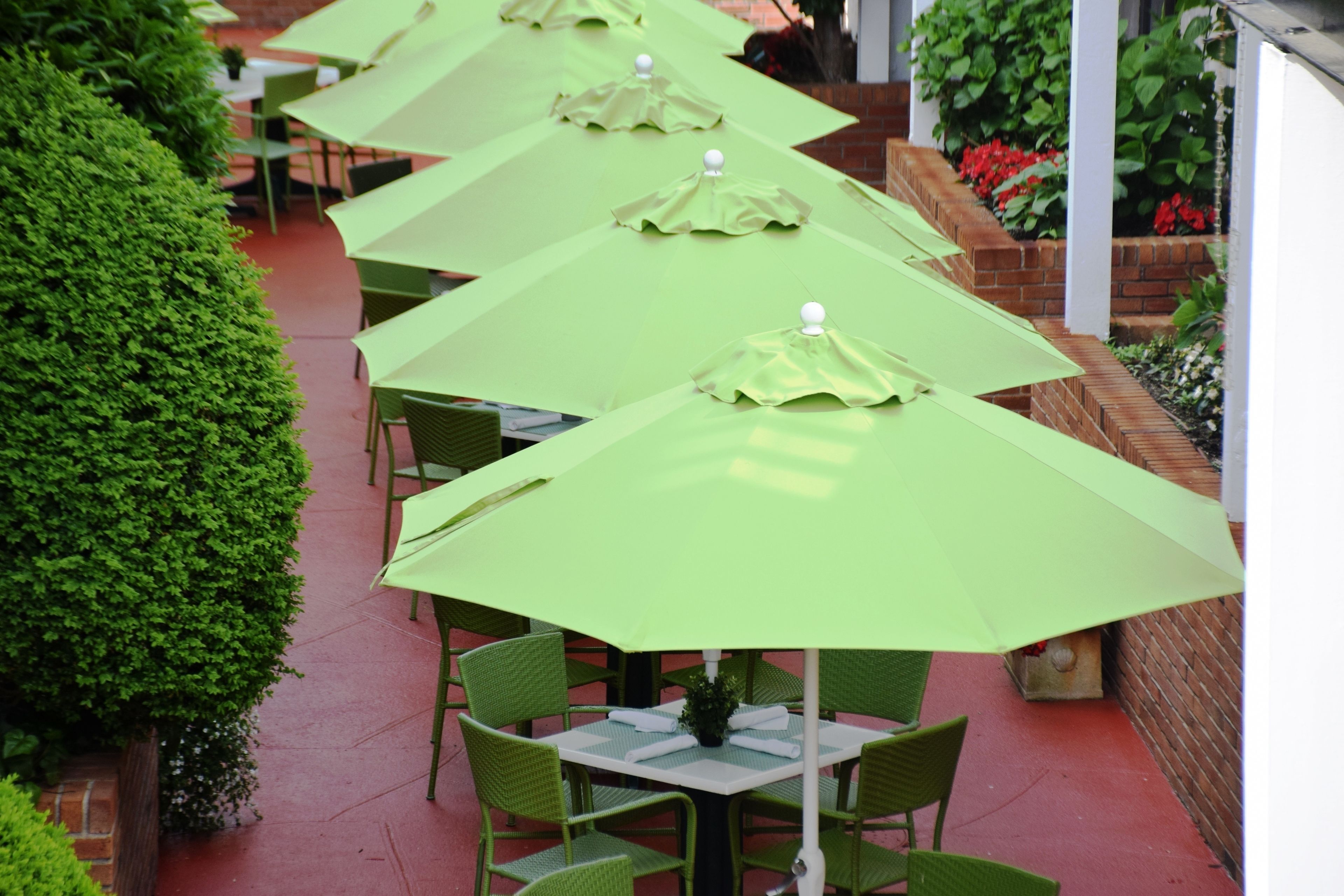 A row of outdoor tables with green umbrellas, surrounded by brick walls and greenery.