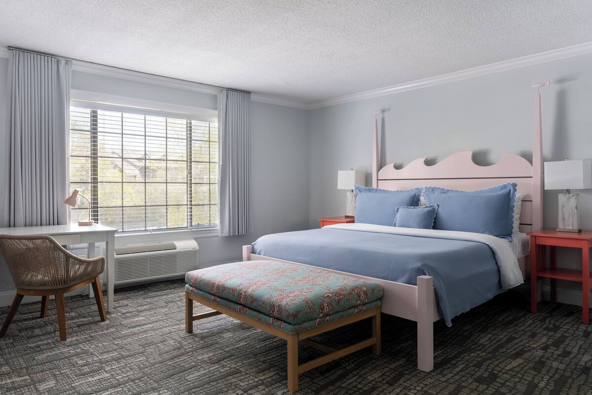 Un dormitorio con cama, escritorio, silla, banco y ventana con cortinas.