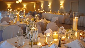 Une salle de banquet avec des tables rondes dressées pour le repas, chacune agrémentée de bougies, de verres et de compositions florales.