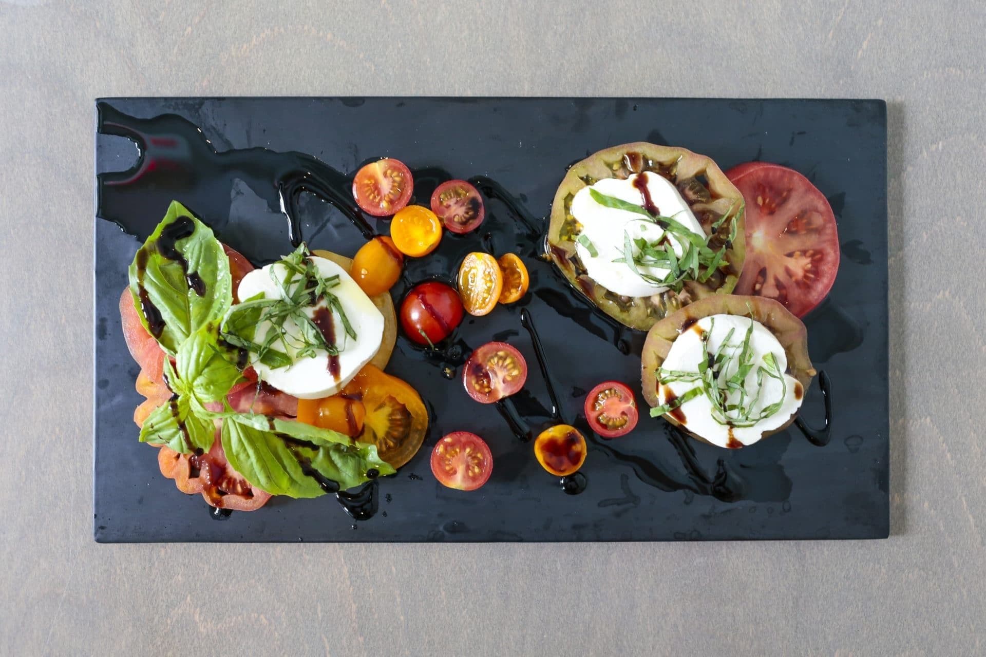 A rectangular black slate serving platter with a variety of food items including tomatoes, mozzarella, and basil leaves.