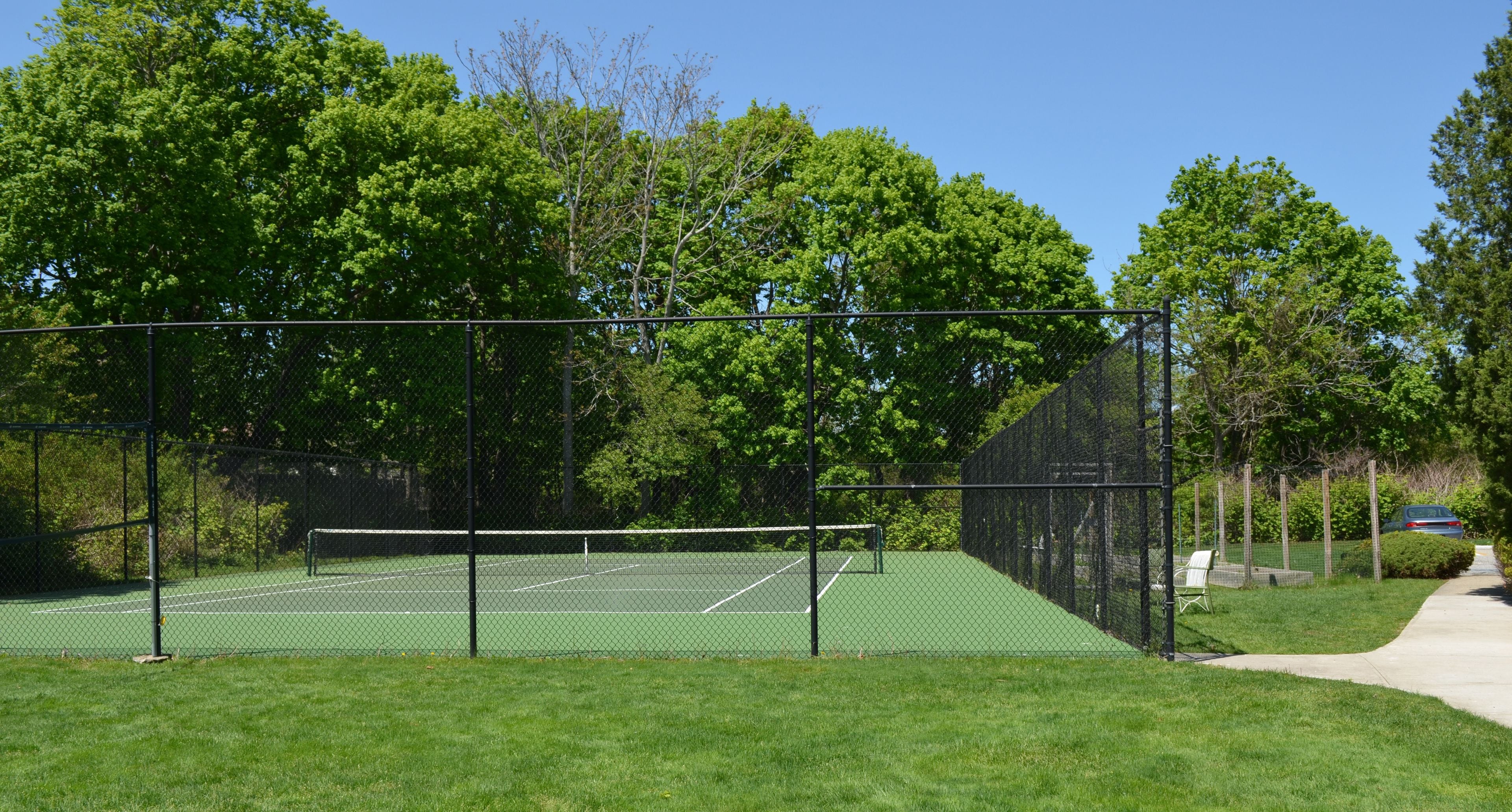 A tennis court surrounded by a chain-link fence, with trees and a pathway in the background.