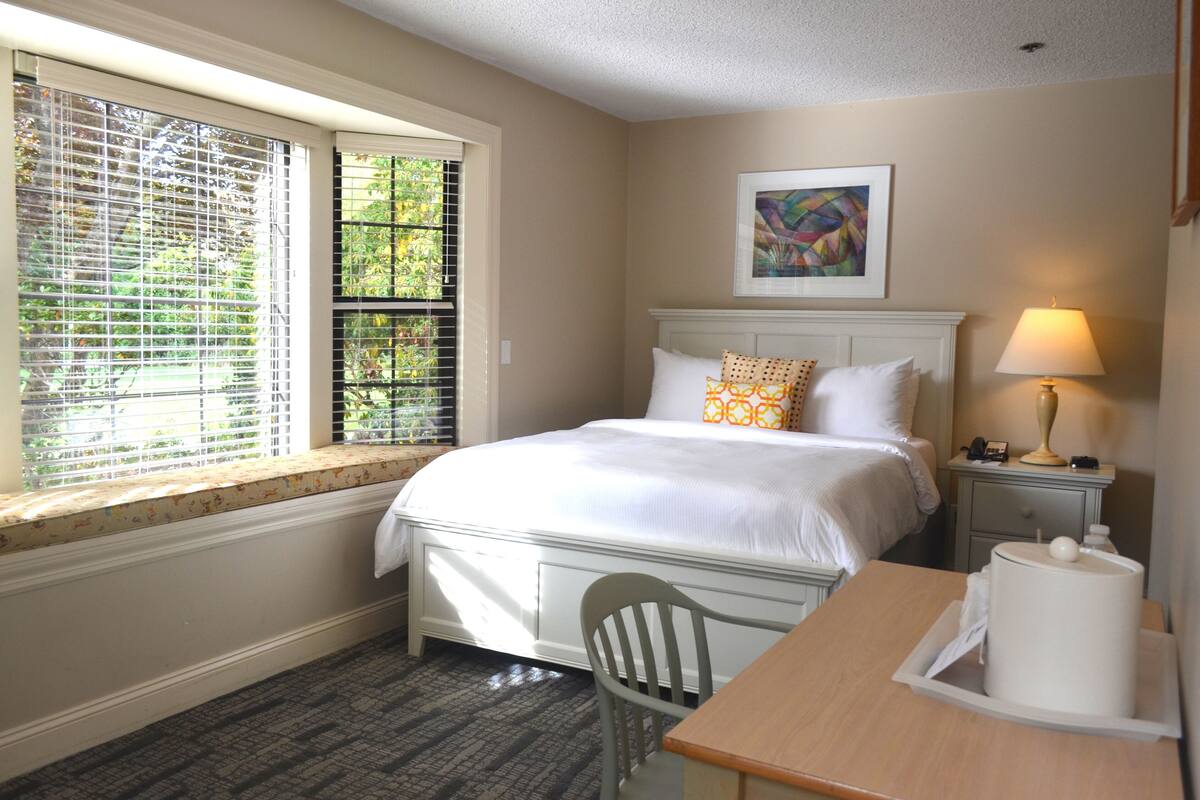 Una habitación de hotel con cama, escritorio, silla, mesita de noche con lámpara y ventana con persianas.