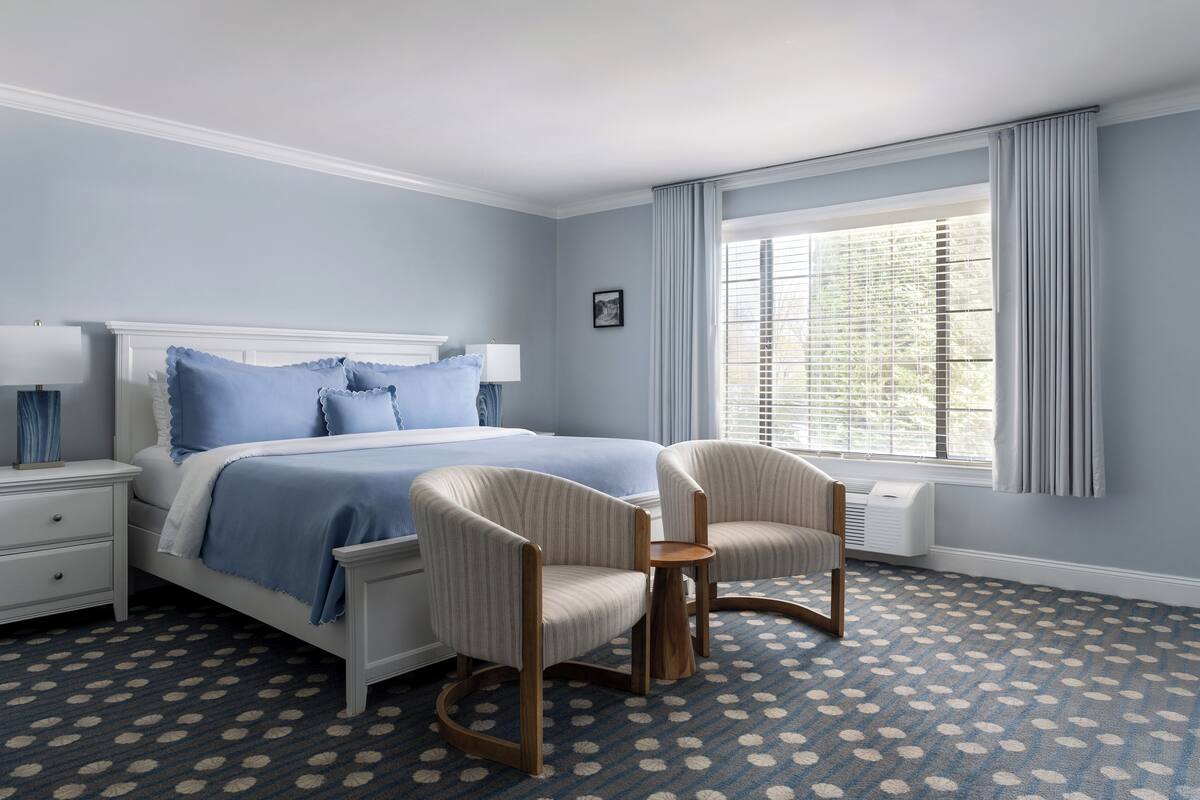 Un dormitorio con cama, dos sillas, mesita de noche, lámpara y ventana con cortinas.