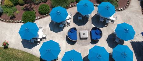 An aerial view of a garden with multiple seating areas under blue umbrellas, surrounded by well-maintained greenery and a paved walkway.