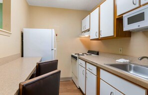 Une cuisine compacte avec des meubles blancs, une cuisinière, un four à micro-ondes, un évier et un petit coin repas avec une chaise.