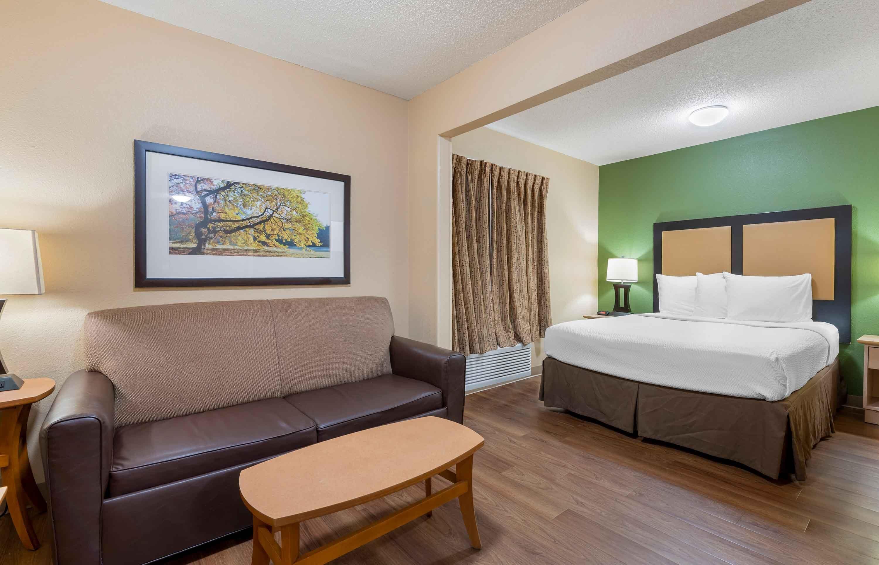 Habitación de hotel con cama, sofá, una mesita, un cuadro en la pared y una ventana con cortinas.