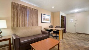 A hotel room with a brown leather sofa, a wooden coffee table, a desk with two chairs, a framed picture on the wall, and a door with a light switch. - Extended Stay America Suites Chicago Lombard Yorktown Center (Lombard)