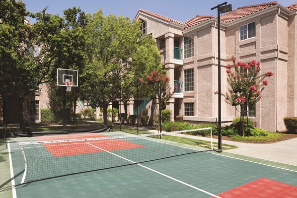 An outdoor tennis court adjacent to a building with a basketball hoop.
