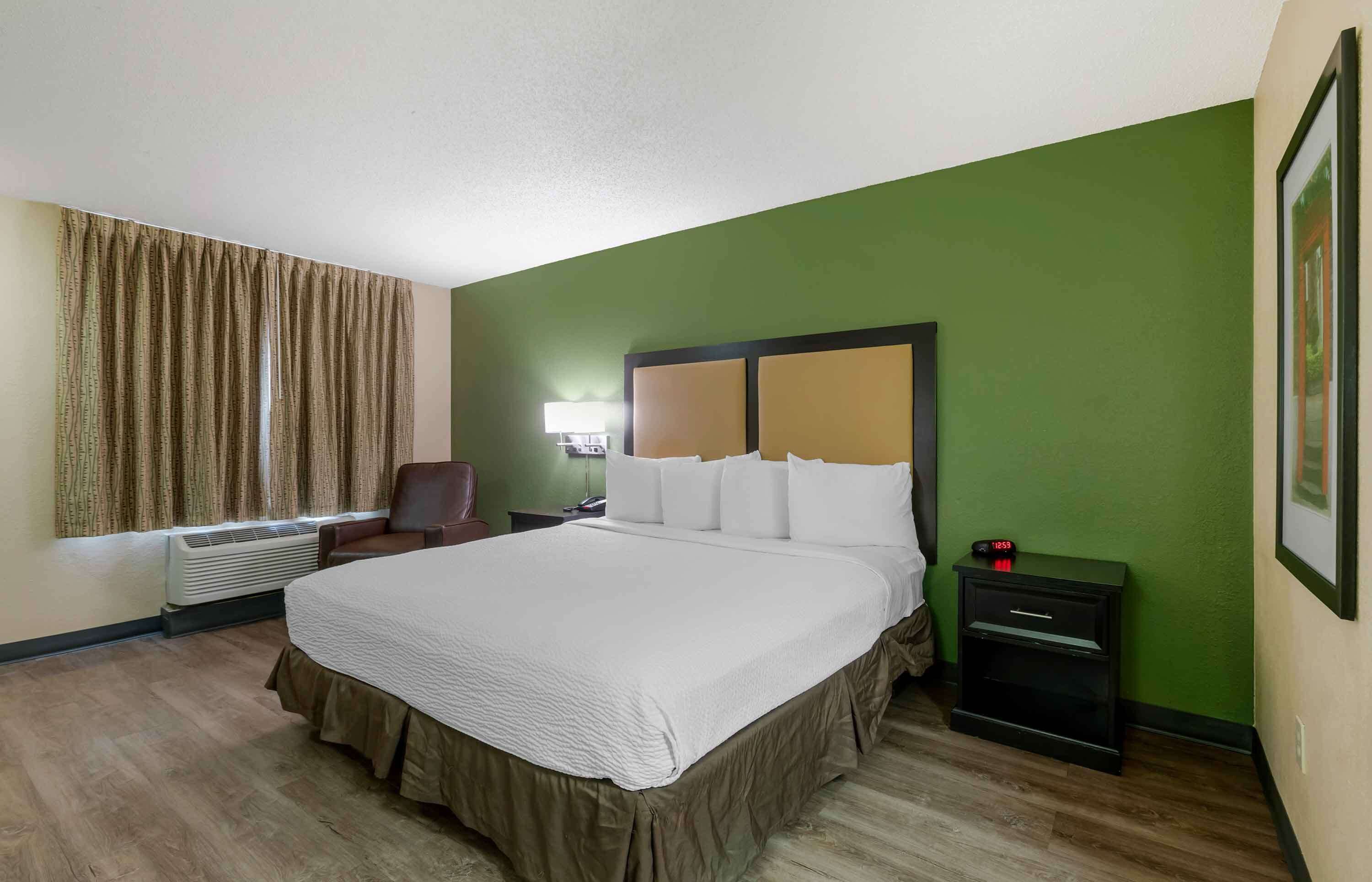 A hotel room with a large bed, a chair, a bedside table with a clock, and a window with curtains.