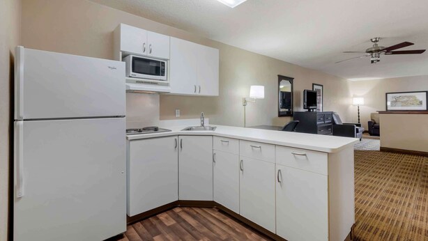 Cocina compacta con gabinetes blancos, microondas y refrigerador. Hay un lavamanos, un ventilador de techo y una pequeña zona de estar con una silla y una mesa.
