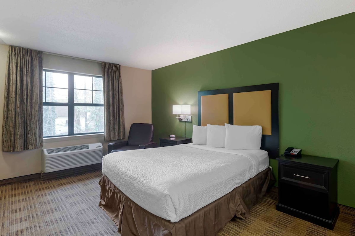 Ein Hotelzimmer mit einem großen Bett, einem Sessel, einem Nachttisch, einer Lampe und einem Fenster mit Vorhängen.