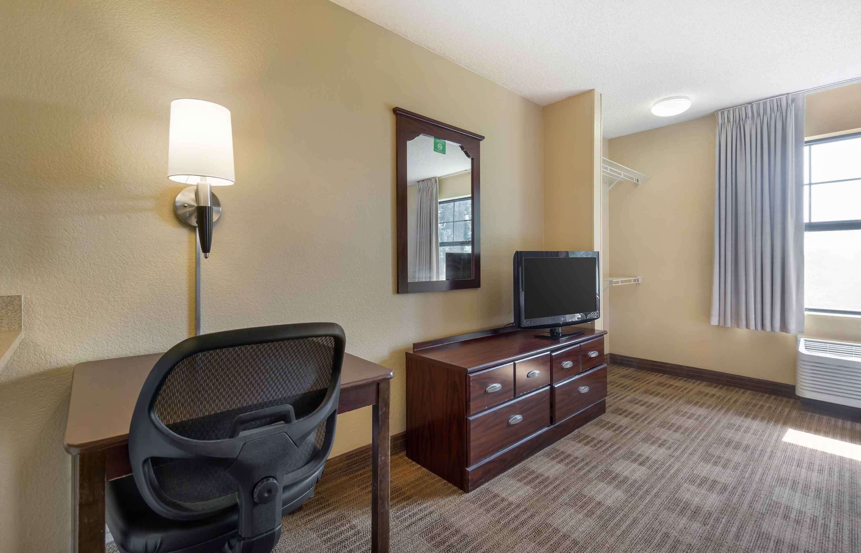 Habitación de hotel con escritorio, una silla, televisor sobre un mueble de madera, un espejo en la pared y ventana con cortinas.