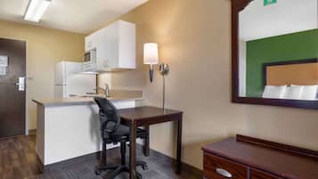 A hotel room with a kitchenette, a desk with a chair, a bedside table, and a mirror.