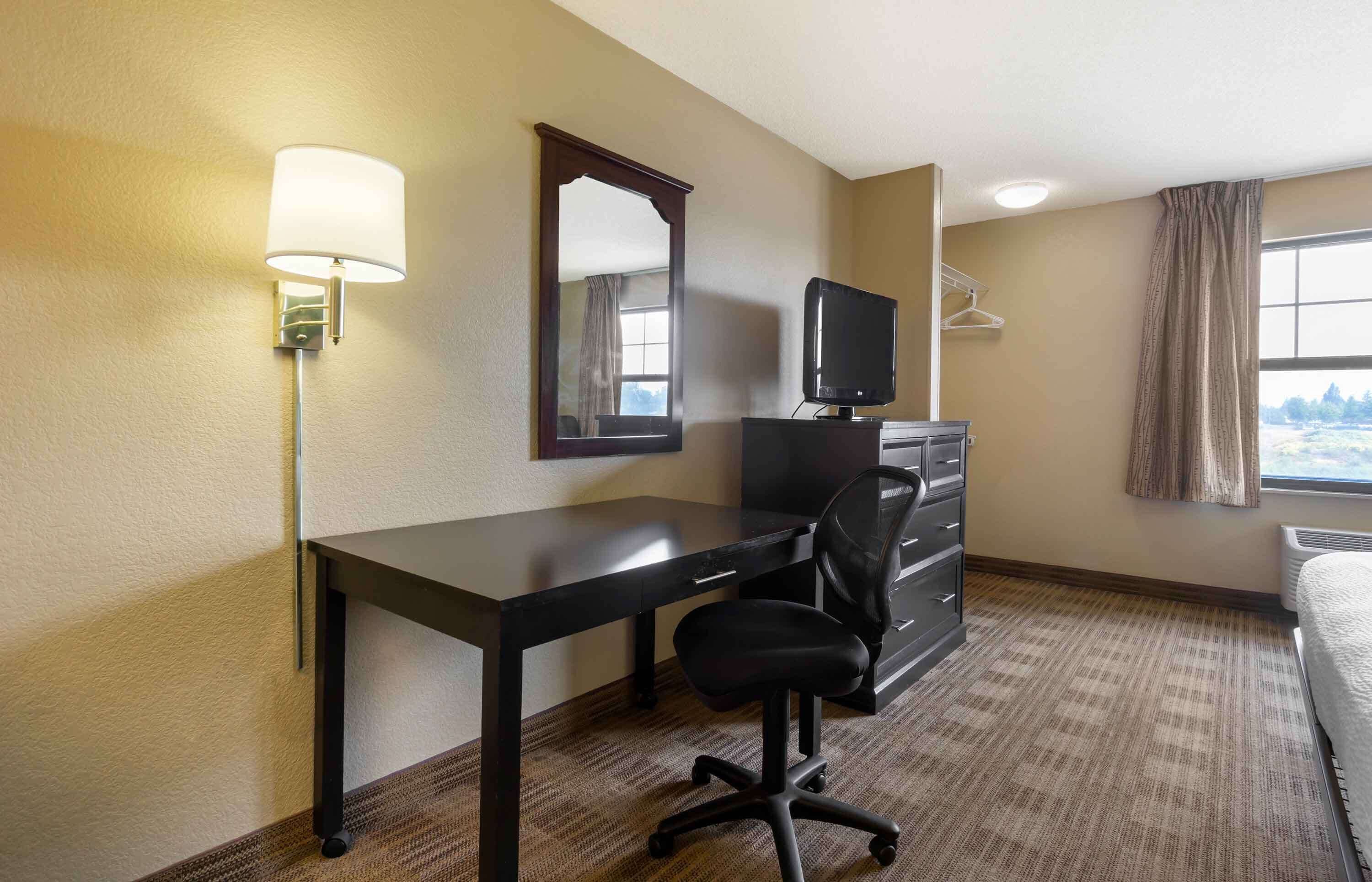 A hotel room with a desk, chair, dresser, TV, and a window with curtains.