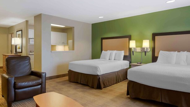 A hotel room with two beds, a chair, a desk, and a kitchenette.