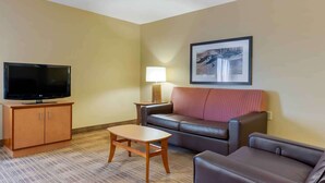 A hotel room with a flat-screen TV on a wooden cabinet, a small wooden table, a leather sofa, a standing lamp, and a framed picture on the wall.