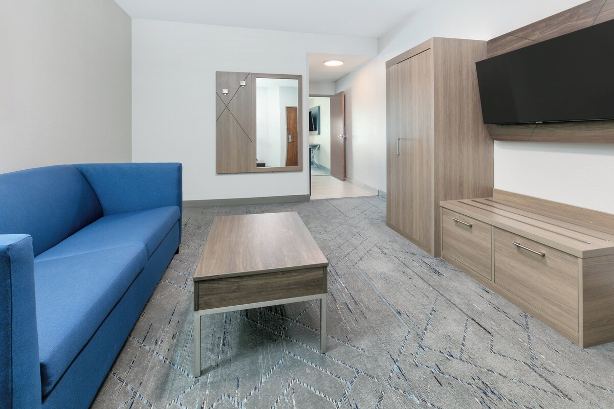 A modern hotel room with a blue sofa, a wooden coffee table, a wardrobe, and a flat-screen TV mounted on the wall.